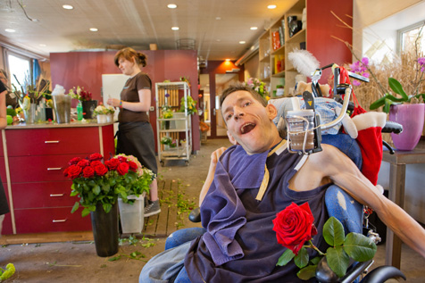 Das Foto zeigt einen Menschen im Rollstuhl mitten im Blumenladen der Stiftung Wagerenhof. Trotz seiner schweren Beeinträchtigung will er überall teilhaben und freut sich über die rote Rose, die er eben geschenkt bekommen hat.